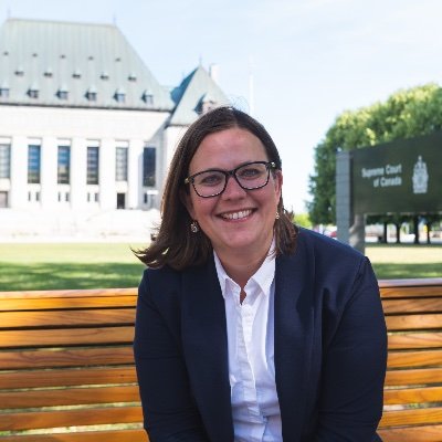 Emilie Taman, a woman with brown hair and glasses wearing a white blouse and navy blazer, sits outside on a bench in front of the Supreme Court of Canada, smiling at the camera.