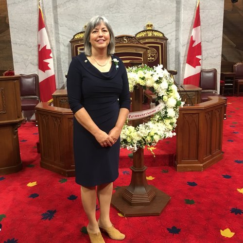 Julia Sneyd, a woman with grey shoulder-length hair wearing a black dress, stands in front of the Senate Speaker's chair in Parliament, smiling at the camera. Behind her is a memorial wreath for her late husband, Paul Dewar