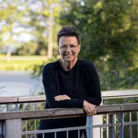 Photo of a smiling Catherine McKenney wearing a black shirt 
                  and leaning on the railing of a footbridge. Behind them is a large 
                  tree and the Ottawa canal